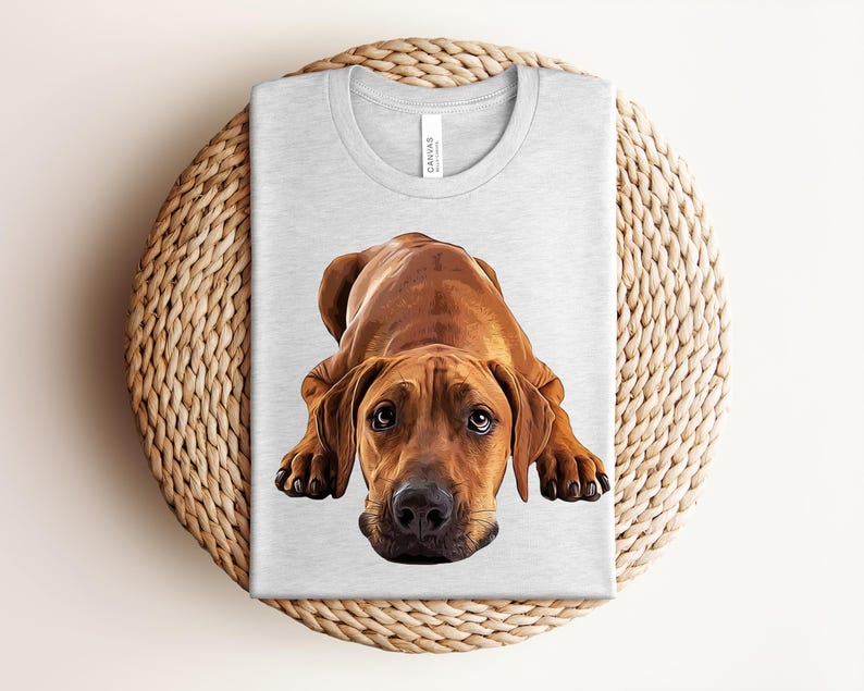 May include: A light gray crew neck t-shirt featuring a detailed, realistic illustration of a brown Rhodesian Ridgeback dog lying down in a prone position, looking directly forward with dark eyes and a black nose, centered on the front of the shirt.