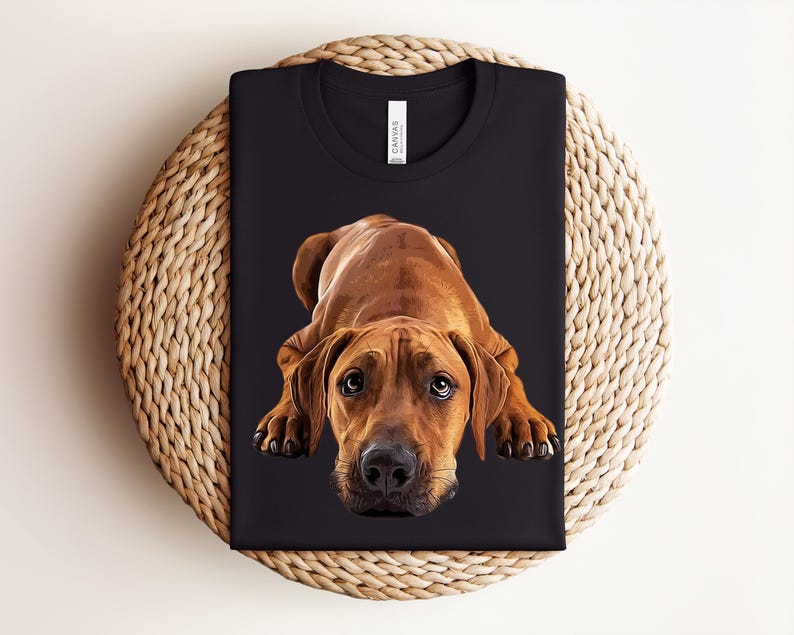 May include: A black crew neck t-shirt featuring a graphic illustration of a brown Rhodesian Ridgeback dog lying down with its head facing forward. The dog has dark eyes and a black nose. The shirt has a white brand label at the inner neckline.