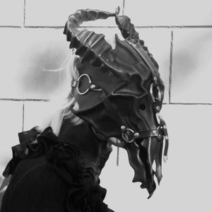 May include: A black leather mask with horns and metal accents. The mask is detailed with a skull-like design and has a strap that goes around the head.