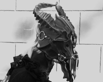 May include: A black leather mask with horns and metal accents. The mask is detailed with a skull-like design and has a strap that goes around the head.