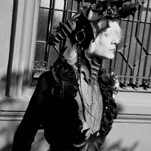 May include: A person wearing a black leather outfit with a dragon-shaped headpiece. The outfit features a corset with a pointed, jagged design and a ruffled collar. The headpiece has a long, flowing tail and is adorned with metal accents.