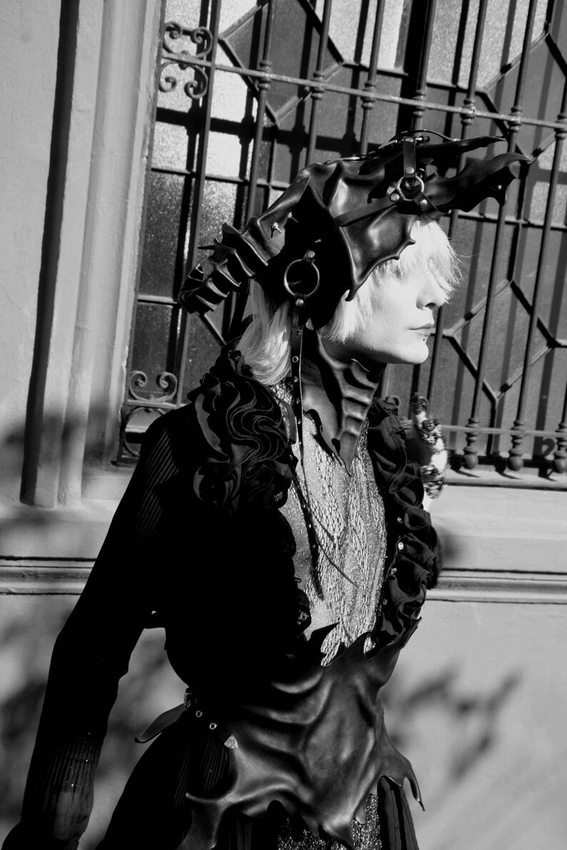 May include: A person wearing a black leather outfit with a dragon-shaped headpiece. The outfit features a corset with a pointed, jagged design and a ruffled collar. The headpiece has a long, flowing tail and is adorned with metal accents.