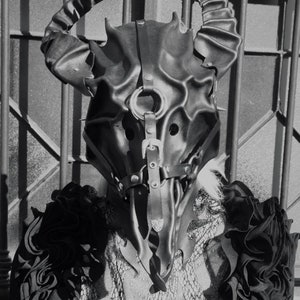 May include: A black leather mask with horns and straps. The mask has a detailed design and is worn by a person with a black and white patterned shirt.