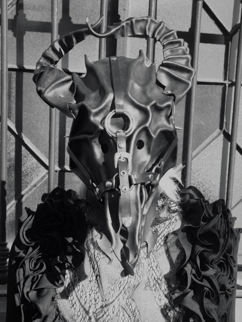 May include: A black leather mask with horns and straps. The mask has a detailed design and is worn by a person with a black and white patterned shirt.