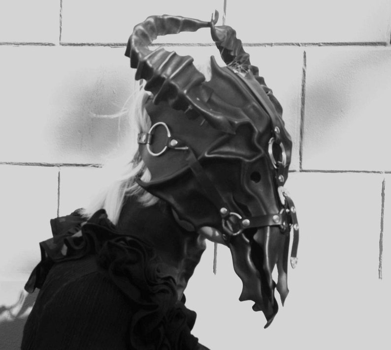 May include: A black leather mask with horns and metal accents. The mask is detailed with a skull-like design and has a strap that goes around the head.
