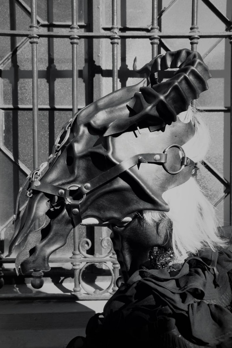 May include: A black leather mask with a dragon design. The mask is worn by a person with white hair. The mask is detailed with straps and buckles.