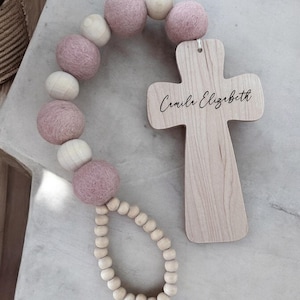 May include: A wooden cross with the name "Camila Elizabeth" inscribed on it, attached to a string of pink felt and wooden beads.