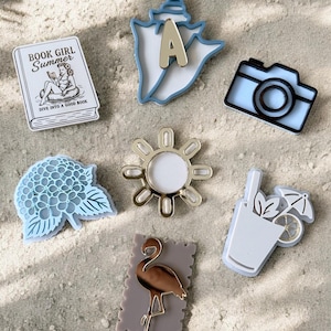 May include: A collection of seven decorative pins featuring a book with the text BOOK GIRL Summer DIVE INTO A GOOD BOOK, a blue seashell with a letter A, a black and white camera, a blue hydrangea, a gold sun, a drink with an umbrella, and a flamingo.