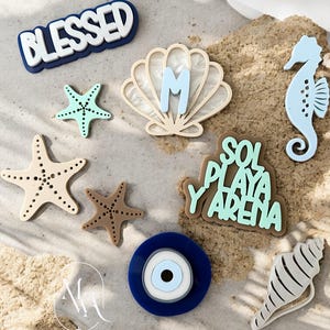 May include: A collection of beach-themed decorative items, including starfish, a seahorse, a seashell, and an evil eye charm. The items are in various colors, including blue, beige, and brown. Some items have text that reads "BLESSED" and "SOL PLAYA Y ARENA".