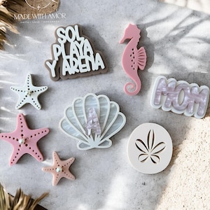 Può includere: Una collezione di ciondoli in acrilico a tema spiaggia in varie forme e colori. Include un cavalluccio marino rosa, stelle marine, una conchiglia con la lettera "A" e ciondoli con le parole "SOL PLAYA Y ARENA" e "MOM".