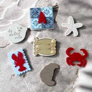 May include: A collection of decorative items, including a red letter "A", a starfish, a crab, a croissant, and a lobster. Other items include a blue tile with a floral design and the words "la dolce vita".