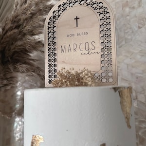 Peut inclure: Une arche en bois avec un motif découpé, comportant le texte "God Bless Marcos" et une croix. L'arche est posée sur une surface blanche avec des accents dorés.