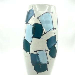 Puede incluir: Un jarrón alto de cerámica blanquecina con forma rectangular redondeada. El jarrón está decorado con formas abstractas en tonos azules y turquesas, delineadas en negro. El diseño recuerda a un estilo de arte moderno.