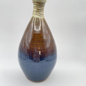 May include: A handcrafted ceramic vase with a teardrop shape. The vase features a gradient of colors, transitioning from a speckled blue base to a rich brown midsection, and a textured tan neck. The vase is approximately 10 inches tall.