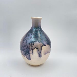 Puede incluir: Un jarrón de cerámica hecho a mano con un cuerpo redondeado y un cuello estrecho. El jarrón presenta un diseño único con una mezcla de colores, incluyendo crema, azul y morado. La superficie tiene un patrón texturizado similar al craquelado.