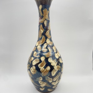 Puede incluir: Un jarrón de cerámica alto, con una base azul oscuro y un cuello delgado. El jarrón está adornado con un patrón de pinceladas de color beige y crema. El borde es de color marrón oscuro. Este jarrón decorativo es adecuado para la decoración del hogar.