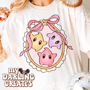 Puede incluir: Sudadera color crema con un dise&ntilde;o caprichoso de tres personajes en forma de estrella en amarillo, morado y rosa, dentro de un marco decorativo con un lazo rosa. El texto "my DARLING CREATES" est&aacute; impreso en la parte inferior.