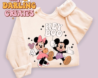 May include: A beige sweatshirt with a Mickey Mouse and Minnie Mouse graphic. The graphic features Mickey and Minnie holding a pumpkin and a lollipop. The text "Hey Boo" is printed above the graphic.