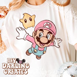Puede incluir: Sudadera de color crema con una ilustraci&oacute;n de dibujos animados de Mario con gorro y peto rosas, y una estrella amarilla. El texto "My Darling Creates" est&aacute; impreso en la parte inferior en una fuente marr&oacute;n y juguetona.