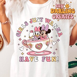 以下が含まれることがあります： 白地にピンクと白のグラフィックがプリントされたTシャツ。ミッキーマウスとデイジーダックの2人の漫画キャラクターがティーカップに座っている。ティーカップの下には「Girls Just Wanna Have Fun!」の文字がピンクと白の文字でプリントされている。Tシャツの右上には「my DARLING CREATES」の文字がレトロなフォントでプリントされている。