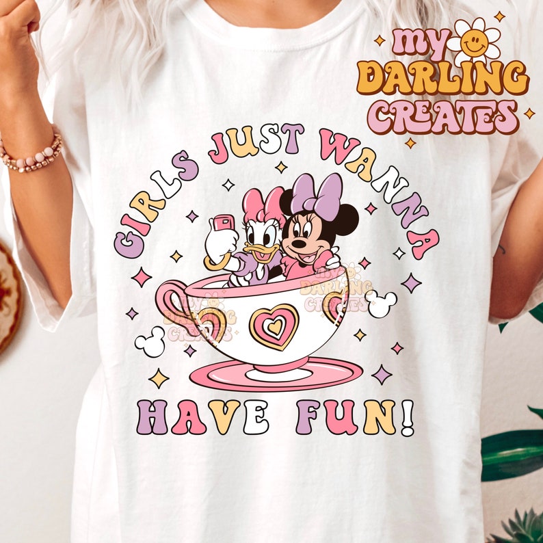 以下が含まれることがあります： 白地にピンクと白のグラフィックがプリントされたTシャツ。ミッキーマウスとデイジーダックの2人の漫画キャラクターがティーカップに座っている。ティーカップの下には「Girls Just Wanna Have Fun!」の文字がピンクと白の文字でプリントされている。Tシャツの右上には「my DARLING CREATES」の文字がレトロなフォントでプリントされている。