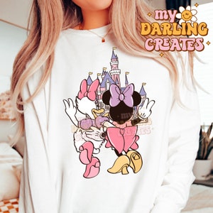 Peut inclure: Sweat-shirt blanc avec un motif de Minnie Mouse et Daisy Duck devant un château rose et violet. Le texte "my darling creates" est dans le coin supérieur droit de l'image.
