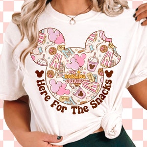 May include: White t-shirt featuring a pink and brown Mickey Mouse head design. The design is filled with various images of snacks, including popcorn, ice cream, and Mickey Mouse ears. The text "Here For The Snacks" is written below the Mickey Mouse head.