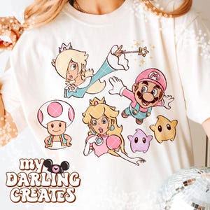 May include: Cream-colored t-shirt featuring cartoon characters from a popular video game. The design includes Mario, Princess Peach, Rosalina, Toad, and other characters. The text "MY DARLING CREATES" is printed at the bottom of the design.