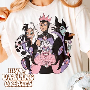 Könnte beinhalten: Weißes T-Shirt mit einem Grafikdruck von vier Disney-Schurken: Ursula, Maleficent, Cruella de Vil und die böse Königin. Die Schurken sind im Cartoon-Stil mit ihren charakteristischen Farben und Accessoires dargestellt. Der Text "my darling creates" ist in einer Retro-Schriftart am unteren Rand des Shirts gedruckt.