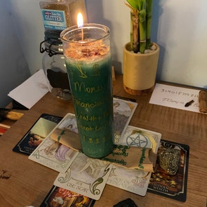 Puede incluir: Una vela verde con letras doradas que dicen "Money, Mancia, Health + Protection" está encendida y rodeada de cartas de tarot, cristales y una pequeña planta en maceta. Las cartas son "The Star", "Ace of Pentacles", y "Sudden Wealth".