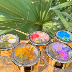 May include: Six glass jars with gold lids, each lid decorated with pressed flowers. The flowers are in various colors, including white, yellow, orange, pink, purple, and blue.