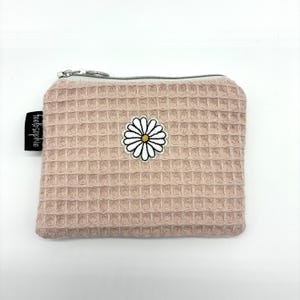 Könnte beinhalten: Eine rosafarbene, strukturierte Geldbörse mit einem Gänseblümchen-Patch. Die Geldbörse hat einen silbernen Reißverschluss und ein kleines schwarzes Etikett mit der Aufschrift "feelosophie". Der Gänseblümchen-Patch ist weiß mit gelber Mitte und schwarzem Umriss.