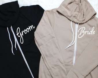 Pode incluir: Duas sweatshirts com capuz, uma preta e outra bege, com zíperes brancos e texto branco. A sweatshirt preta diz "groom" e a sweatshirt bege diz "bride".