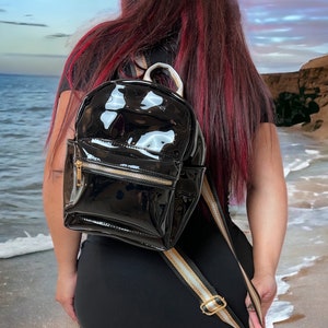 Puede incluir: Una mochila negra de charol con detalles dorados y una correa blanca y dorada. La mochila la lleva puesta una persona con el pelo largo y rojo, de pie en una playa.