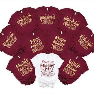 May include: A collection of burgundy t-shirts with gold lettering, including phrases like "Bridesmaid," "Maid of Mischief," and "Mischief Coordinator." One white t-shirt reads "From Mischief to Mrs." with a ring graphic. The shirts are likely for a bridal party.