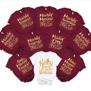 May include: A collection of burgundy and white t-shirts with gold lettering. The burgundy shirts have phrases like "Mischief Supporter" and "Mischief Motivator." One white shirt reads "I Solemnly Swear, It's My 9th Birthday."