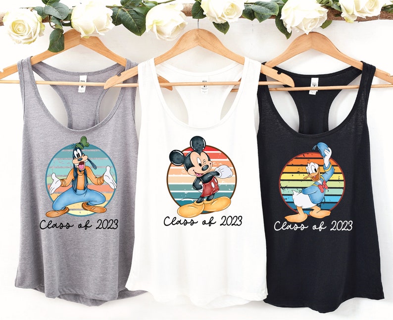 May include: Three tank tops with a retro-style design featuring Mickey Mouse, Donald Duck, and Goofy. Each tank top has a colorful striped circle with the character in the center. The text "Class of 2023" is printed below each character.