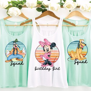 May include: Three mint green tank tops with a retro-style graphic of Disney characters. The center tank top is white and features Minnie Mouse with the text "Birthday Girl". The left and right tank tops feature Goofy and Pluto, respectively, with the text "Squad".