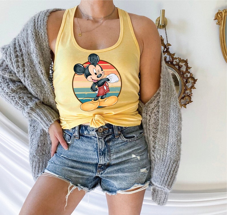 May include: A yellow tank top with a retro-style graphic of Mickey Mouse. The graphic features Mickey Mouse in a red and black outfit, standing in front of a sunset-colored background.