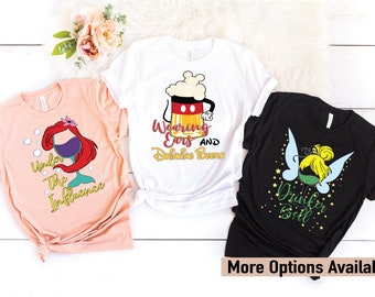 Puede incluir: Tres camisetas gráficas con diseños temáticos de Disney. Una es de color melocotón con Ariel y el texto "Under The Influence". Otra es blanca con un diseño de cerveza de Mickey Mouse y el texto "Wearing Ears and Drinking Beers". La tercera es negra con Campanilla y el texto "Drunker Bell".
