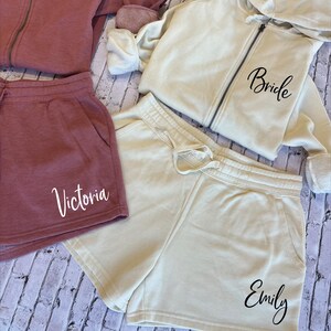 May include: A selection of casual clothing in mauve and cream hues. Includes a mauve zip-up hoodie, shorts with "Victoria", a cream zip-up hoodie with "Bride" and cream shorts with "Emily". The garments are arranged on a light wood-effect surface.