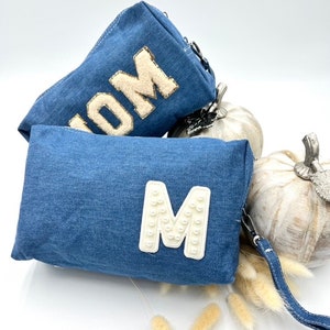 Puede incluir: Dos bolsas de cosméticos de mezclilla con cierres de cremallera. Una bolsa tiene la palabra "MOM" en letras blancas con brillo dorado. La otra bolsa tiene una gran letra blanca "M" hecha de cuentas de perlas.