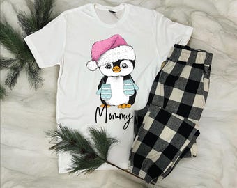 May include: White t-shirt with a penguin graphic wearing a pink Santa hat and blue mittens. The word "Mommy" is written below the penguin. The shirt is paired with black and white plaid pajama pants.