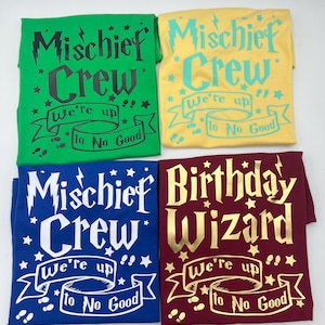 May include: Four folded t-shirts in green, yellow, blue, and burgundy. Each shirt has a different design in a similar style. The green, blue, and yellow shirts say "Mischief Crew" with the phrase "We're up to No Good". The burgundy shirt says "Birthday Wizard".