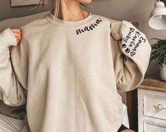 Puede incluir: Una sudadera beige con la palabra "mama" en el hombro izquierdo y el texto "El perro favorito de la abuela" con huellas de patas en la manga derecha.