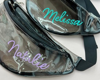May include: Two clear plastic fanny packs with black adjustable straps. The fanny pack on top has the name "Melissa" written in turquoise lettering. The fanny pack on the bottom has the name "Natalie" written in purple lettering.