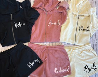 May include: A collection of matching loungewear sets, including black, dusty rose, and cream-colored zip-up hoodies and shorts. The hoodies have the names "Victoria", "Amanda", and "Claudia" printed on them. The shorts are labeled "Morning", "Bridesmaid", and "Bride".