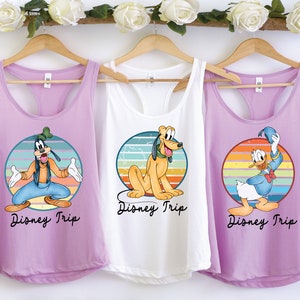 May include: Three purple, white, and light purple tank tops with a retro rainbow design and a Disney character on each. The tank tops say "Disney Trip" on the bottom. The characters are Goofy, Pluto, and Donald Duck.