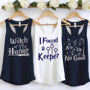 May include: Three tank tops with text and graphics are displayed on wooden hangers. The navy blue tank top says "Witch of Honor" in silver. The white tank top says "I Found a Keeper" in blue. The navy blue tank top says "We Are Up To No Good" in silver.