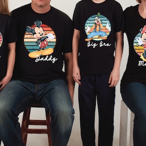 May include: A set of four black t-shirts with a vintage-style design featuring Disney characters. The shirts have the text "Daddy", "Mammy", "Big Bro", and "Mila" printed on them.